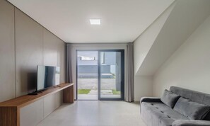 Een moderne woonkamer met een flatscreen televisie op een houten dressoir, een grijze bank en een schuifdeur die uitkomt op een terras.