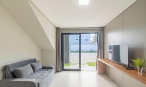 Een moderne woonkamer met een grijze bank, een flatscreen televisie op een houten console, en een schuifdeur die leidt naar een buitenruimte.