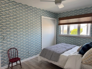 Quarto com papel de parede estampado, cama com manta listrada, cadeira de madeira e janela com vista para vegetação.