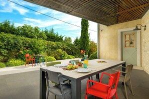 Een terras met eettafel voor twee personen, omringd door stoelen, met uitzicht op een tuin met diverse planten en een hoge boom.