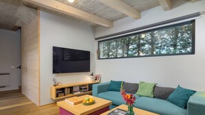 Ein modernes Wohnzimmer mit einem an der Wand montierten Flachbildfernseher, einem petrolfarbenen Sofa mit vielen Kissen, einem Holztisch mit einer Vase und Büchern sowie einem Fenster mit Blick auf Bäume.