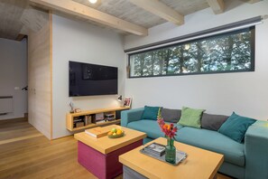 A modern living room with a flat-screen TV mounted on the wall, a teal sofa with multiple cushions, a wooden coffee table with a vase and books, and a window with a view of trees. - Villa Casa LaBobina (Istria - Kurili) (Kurili)