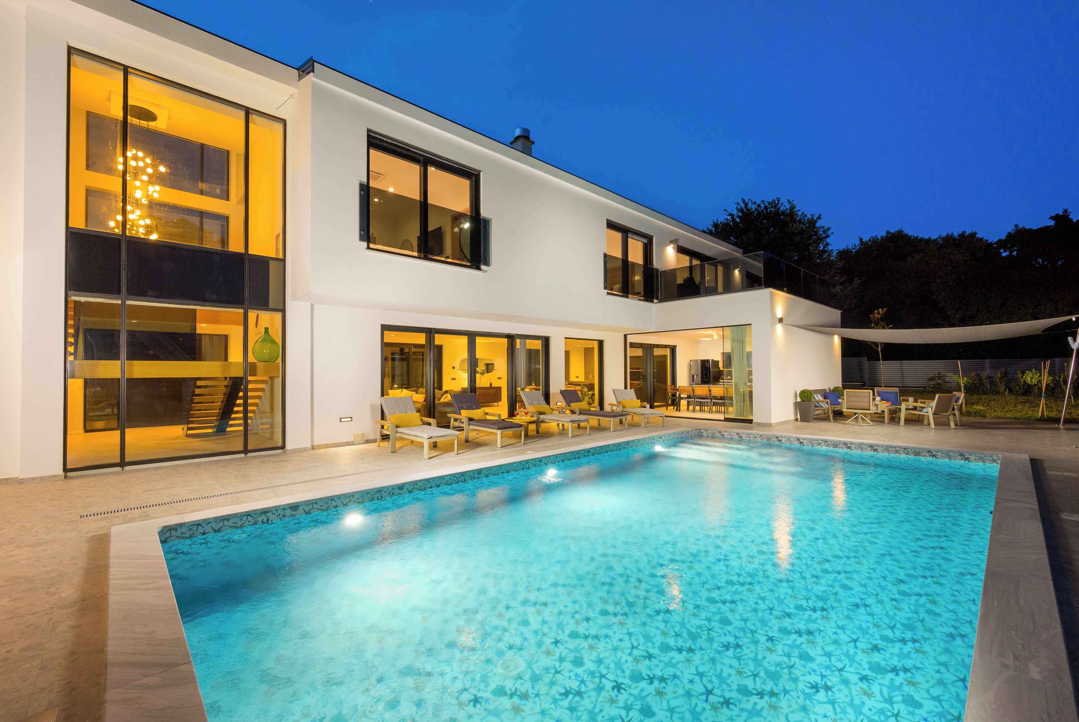 Et moderne hus med to etager, en swimmingpool, store vinduer og udendørs møbler.