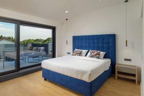 Ein modernes Schlafzimmer mit einem großen Bett, einer Glastür, die auf einen Balkon führt, und Blick ins Grüne.