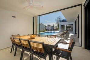 Un espace repas moderne avec vue sur la piscine, comprenant une table et des chaises en bois, ainsi qu’un ventilateur de plafond.