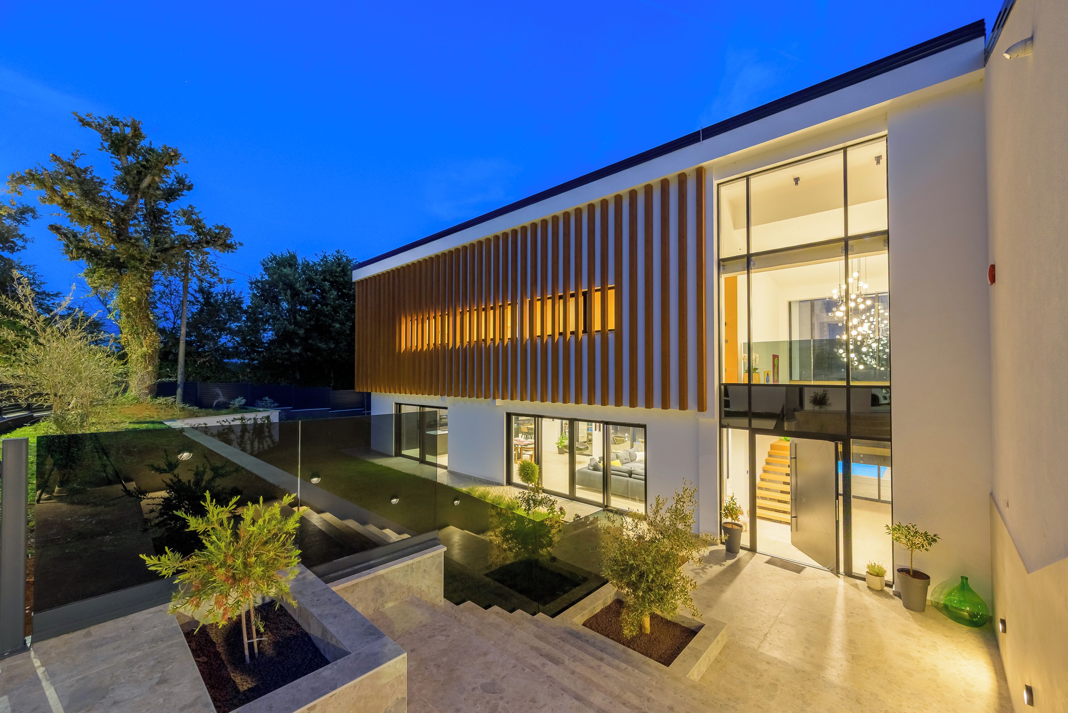 Une maison moderne à deux étages, dotée d’une façade en verre, d’un espace extérieur aménagé et d’un escalier menant à l’étage supérieur.