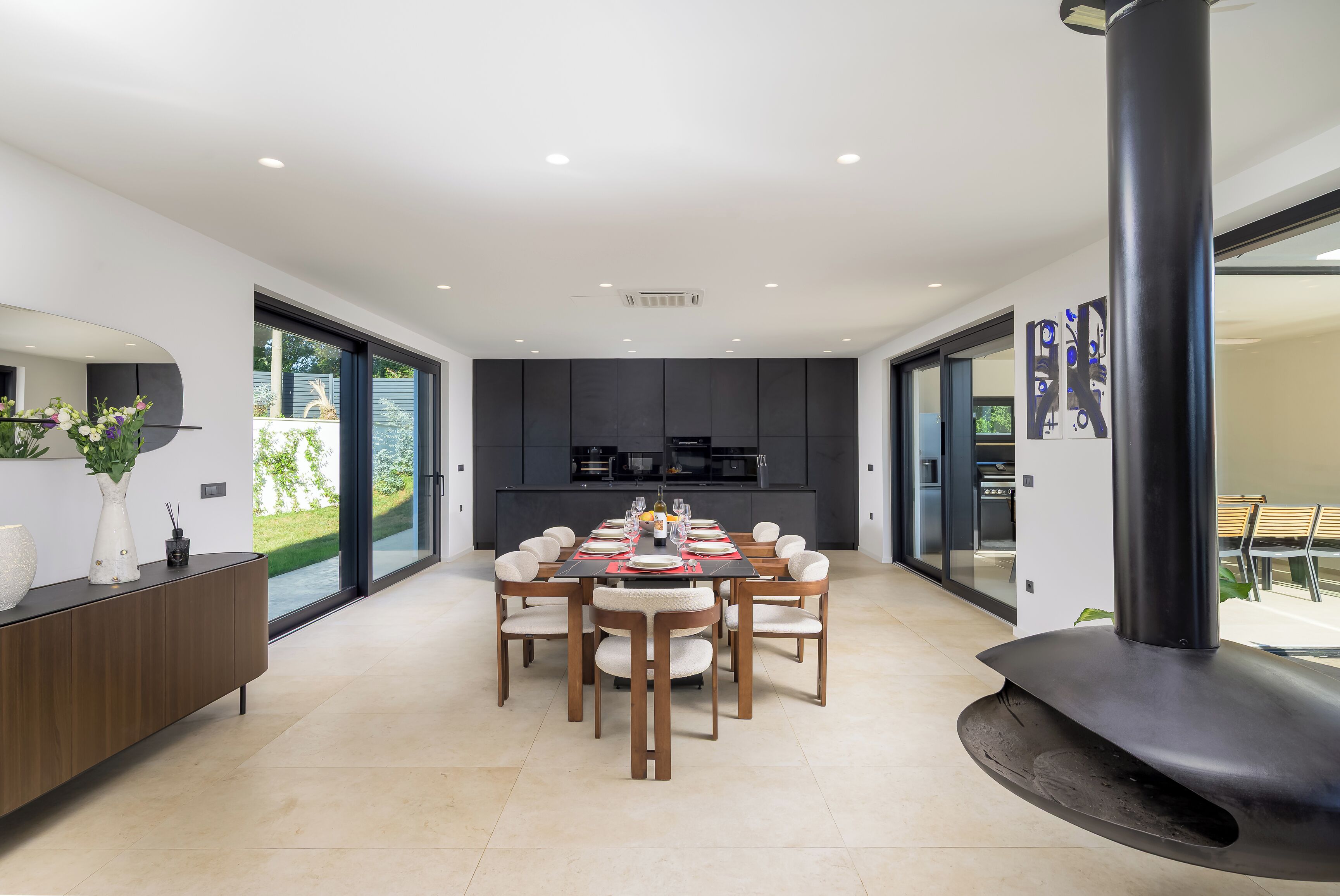 A modern kitchen with dark cabinetry, a dining area with a round table and chairs, and a large window providing a view of the outdoors.