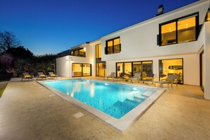 Ein modernes, zweistöckiges Haus mit Swimmingpool, Sitzgelegenheiten im Freien und großen Fenstern.