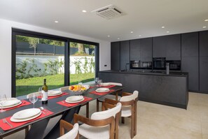 Une cuisine moderne aux meubles sombres, un coin repas avec une table dressée pour quatre personnes, et une vue sur un jardin verdoyant par de grandes fenêtres.