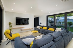 Ein modernes Wohnzimmer mit einem grauen Ecksofa, gelben Akzentstühlen, einem flachbildfernseher an der Wand und einer großen Glasschiebetür, die in einen Garten führt.