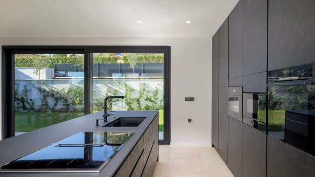 Cocina moderna con una isla grande, electrodomésticos integrados y un ventanal corredero que da a un jardín.