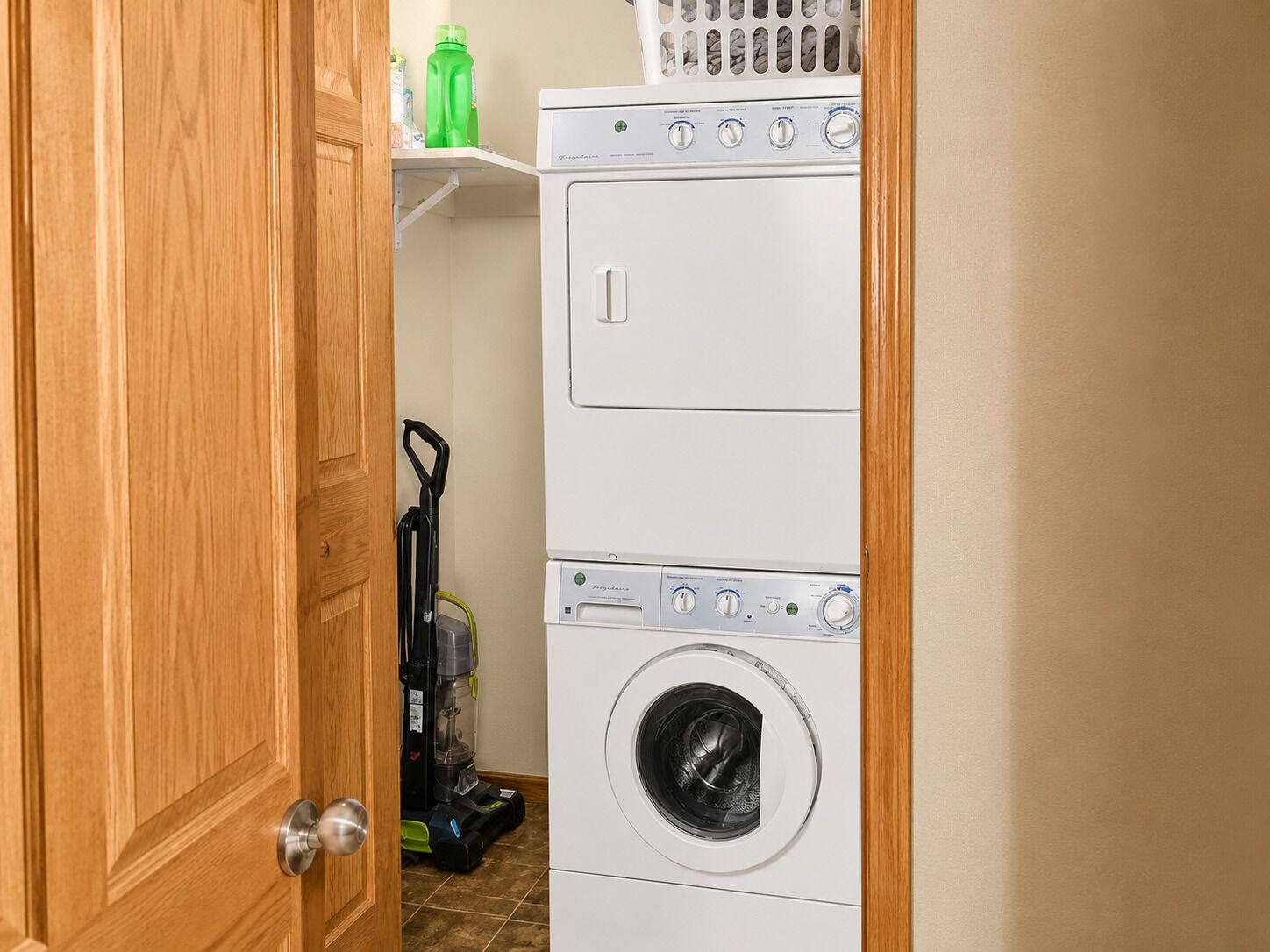 A laundry room with stacked washers and dryers, a vacuum cleaner, and a green bottle on a shelf.