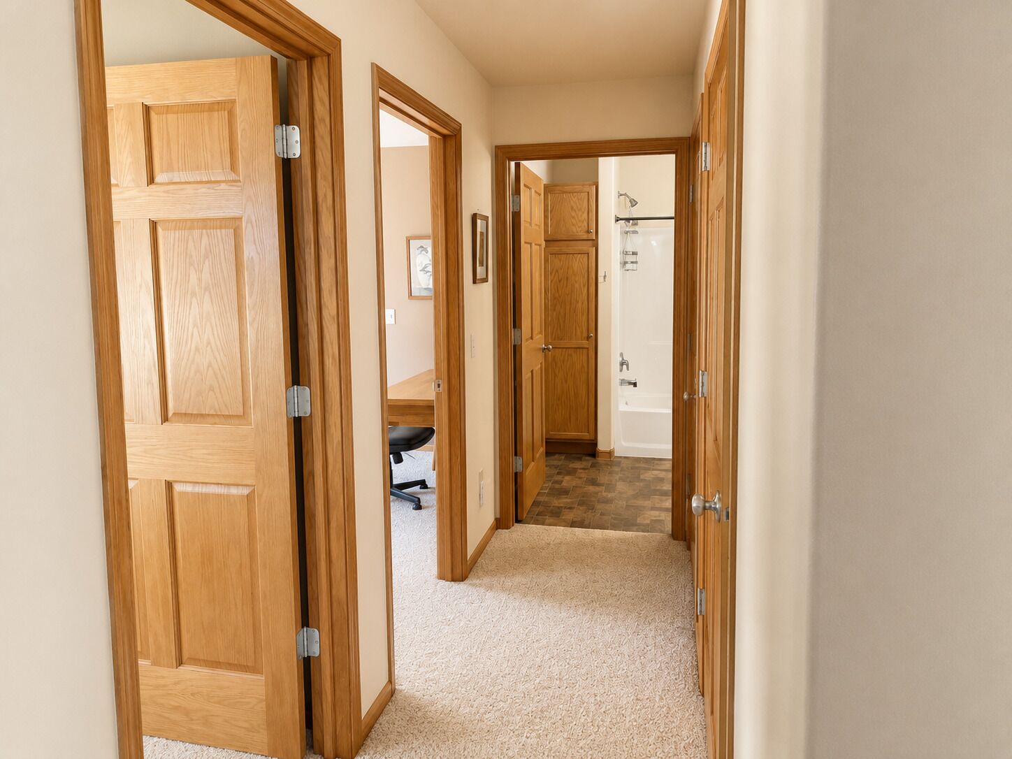 A hallway with wooden doors, carpeted flooring, and a visible bathroom with a bathtub.