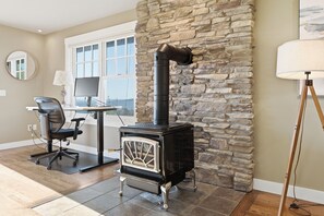 A room with a stone wall, a black wood-burning stove, a desk with a computer, and a chair. - Perch - Studio Retreat | Private Balcony | Sunset Views (Coupeville)