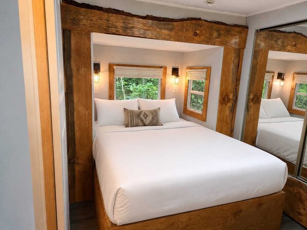 A bed in a room with wooden walls and windows, a mirror on one wall, and a view of greenery outside.