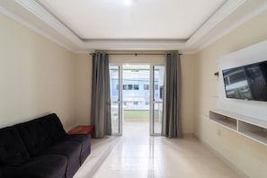 A living room with a black sofa, a flat-screen TV mounted on the wall, and a sliding glass door leading to an outdoor balcony. - Apt With Balcony & BBQ 78m From the Beach Obs0101 (Itapema)