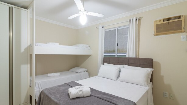 A hotel room with a single bed, a smaller bed on the left, a ceiling fan, a window with curtains, and a wall-mounted air conditioning unit. - Apt With Balcony & BBQ 78m From the Beach Obs0101 (Itapema)
