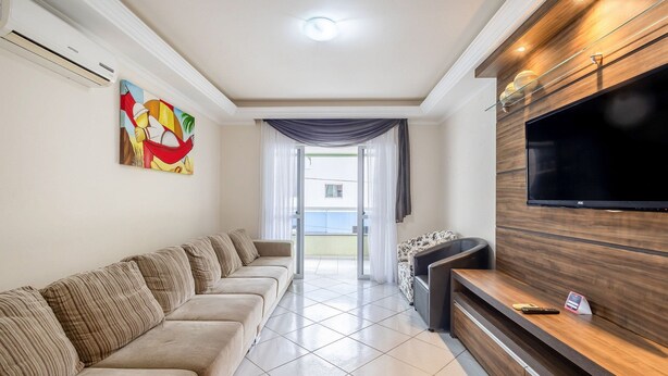 A modern living room with a beige sofa, a flat-screen TV, and a wooden entertainment unit. - Apt With Balcony Steps From the Beach - Obs0201 (Itapema)