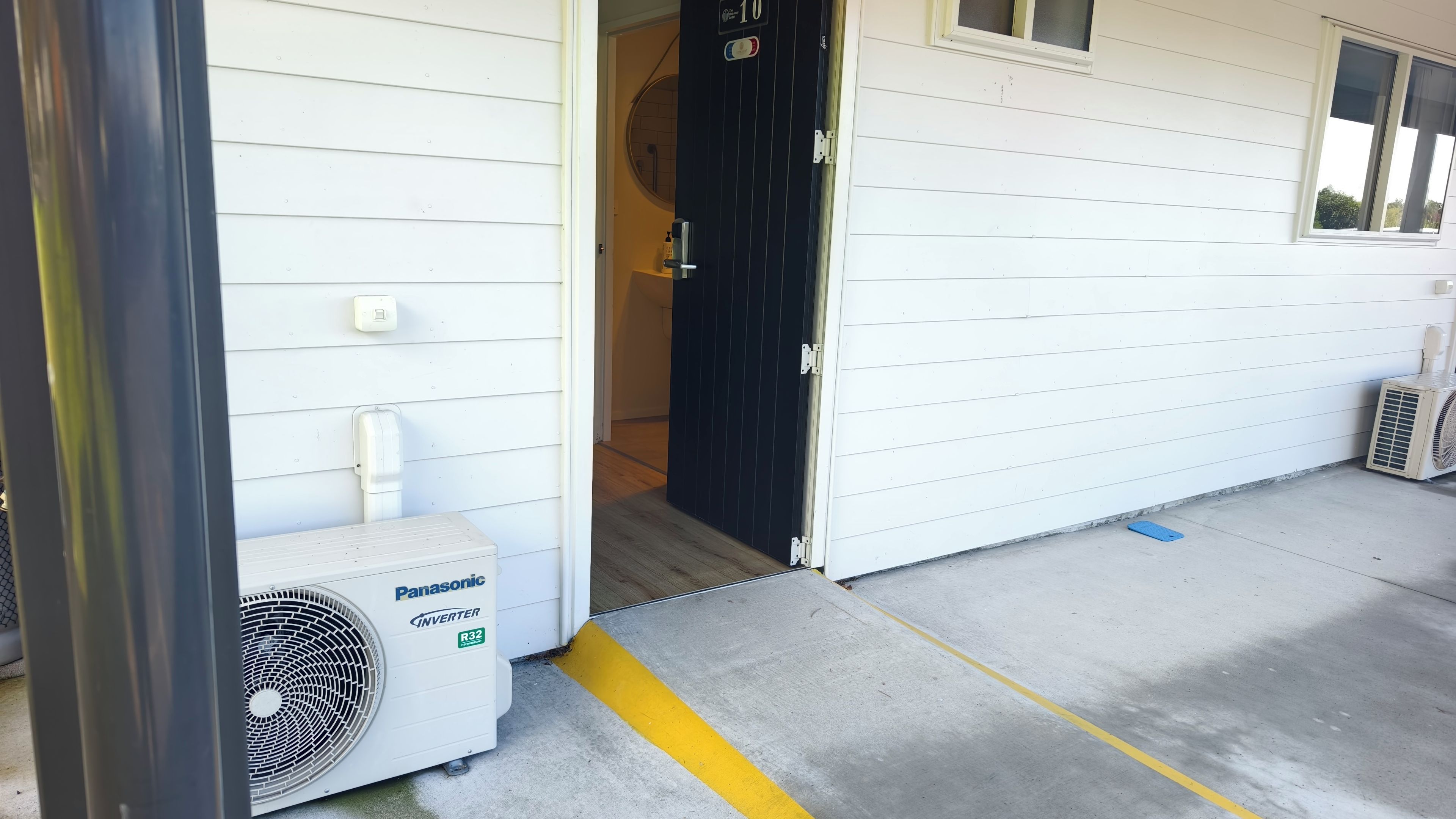 Entrada de un edificio con una puerta negra, una unidad de aire acondicionado blanca con la etiqueta 'Panasonic Inverter R32' y un piso de concreto con líneas amarillas.