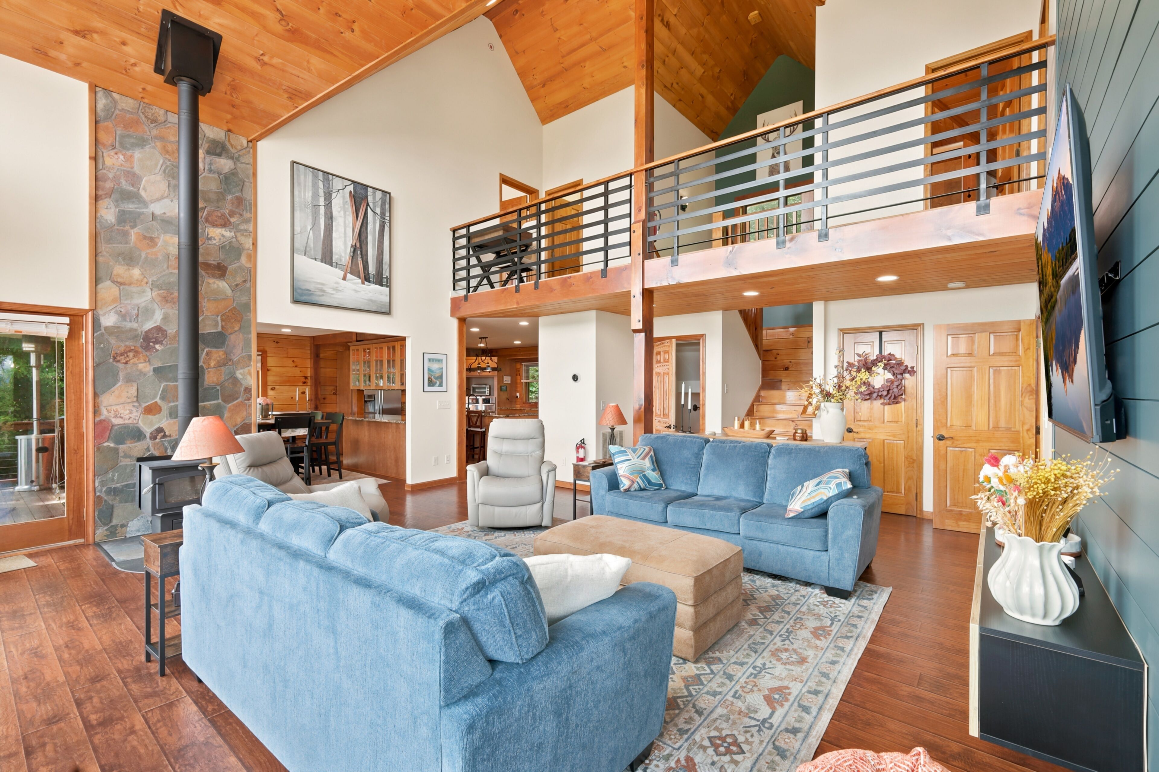 A spacious living area with a high ceiling, a stone fireplace, a dining area, and a kitchen.