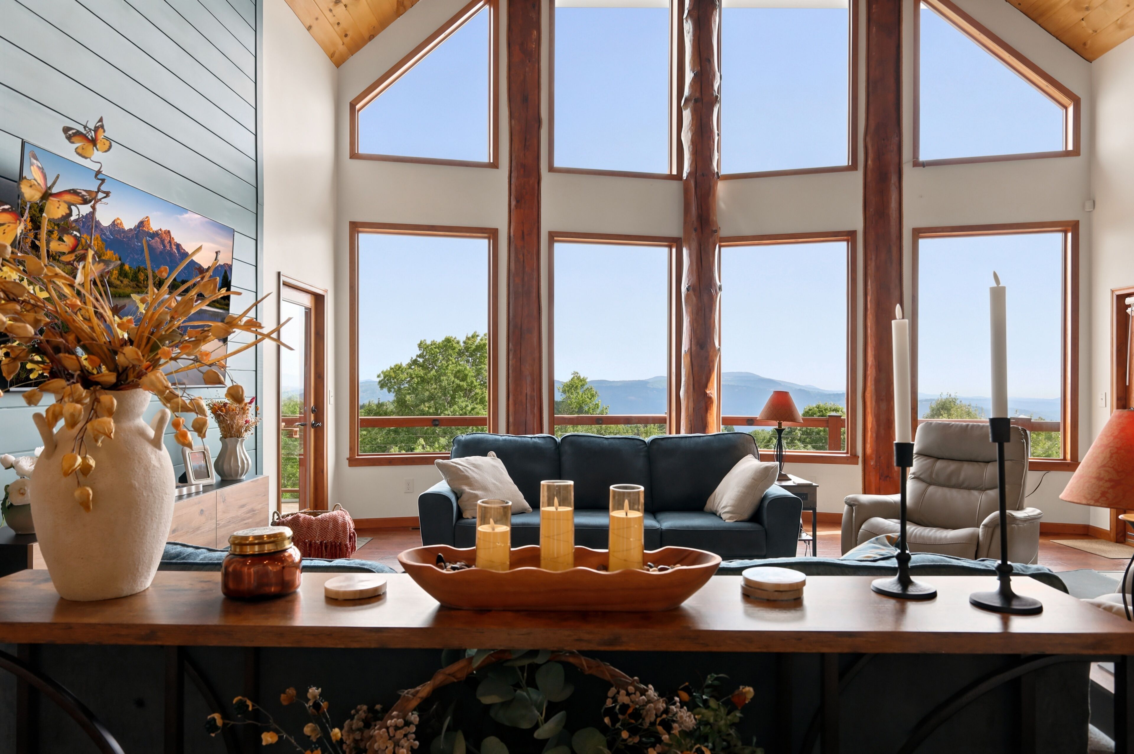 A room with a large window, a blue sofa, a wooden table with candles, and a vase with dried flowers.