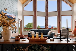 Ein Zimmer mit einem großen Fenster, einem blauen Sofa, einem Holztisch mit Kerzen und einer Vase mit getrockneten Blumen.