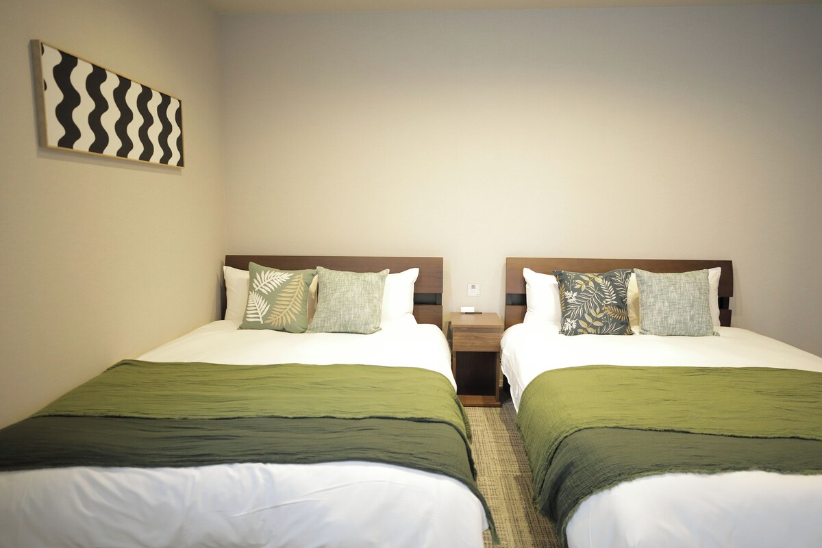 Ein Hotelzimmer mit zwei Einzelbetten, jedes mit einer grünen Bettdecke und weißen Kissen. Dazwischen steht ein kleiner Nachttisch aus Holz mit einer Fernbedienung. Die Wände sind schlicht gehalten, über dem linken Bett hängt ein abstraktes Kunstwerk.