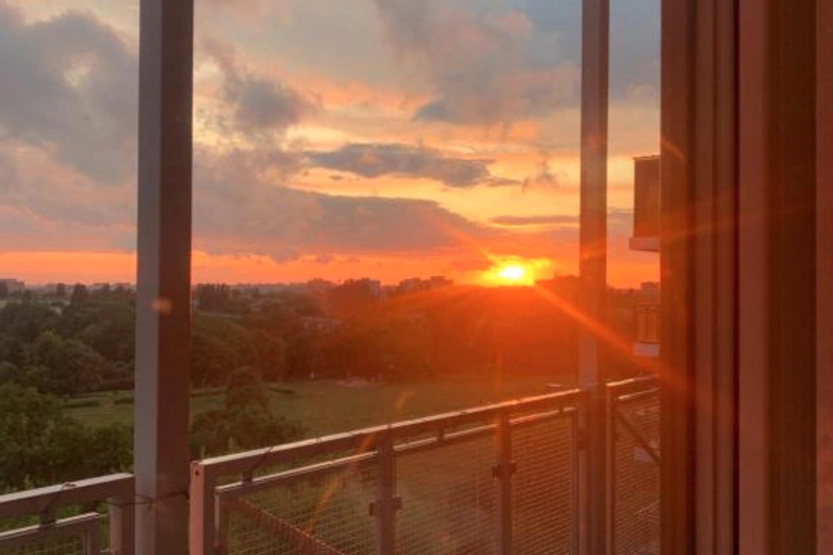 Une vue sur le coucher de soleil depuis un balcon doté d’une balustrade en métal, donnant sur un paysage verdoyant et sur les contours d’une ville.