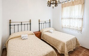 A room with two single beds, a wooden nightstand, a chandelier, and a window with a patterned curtain. - Amazing home in Iznajar (Iznajar)