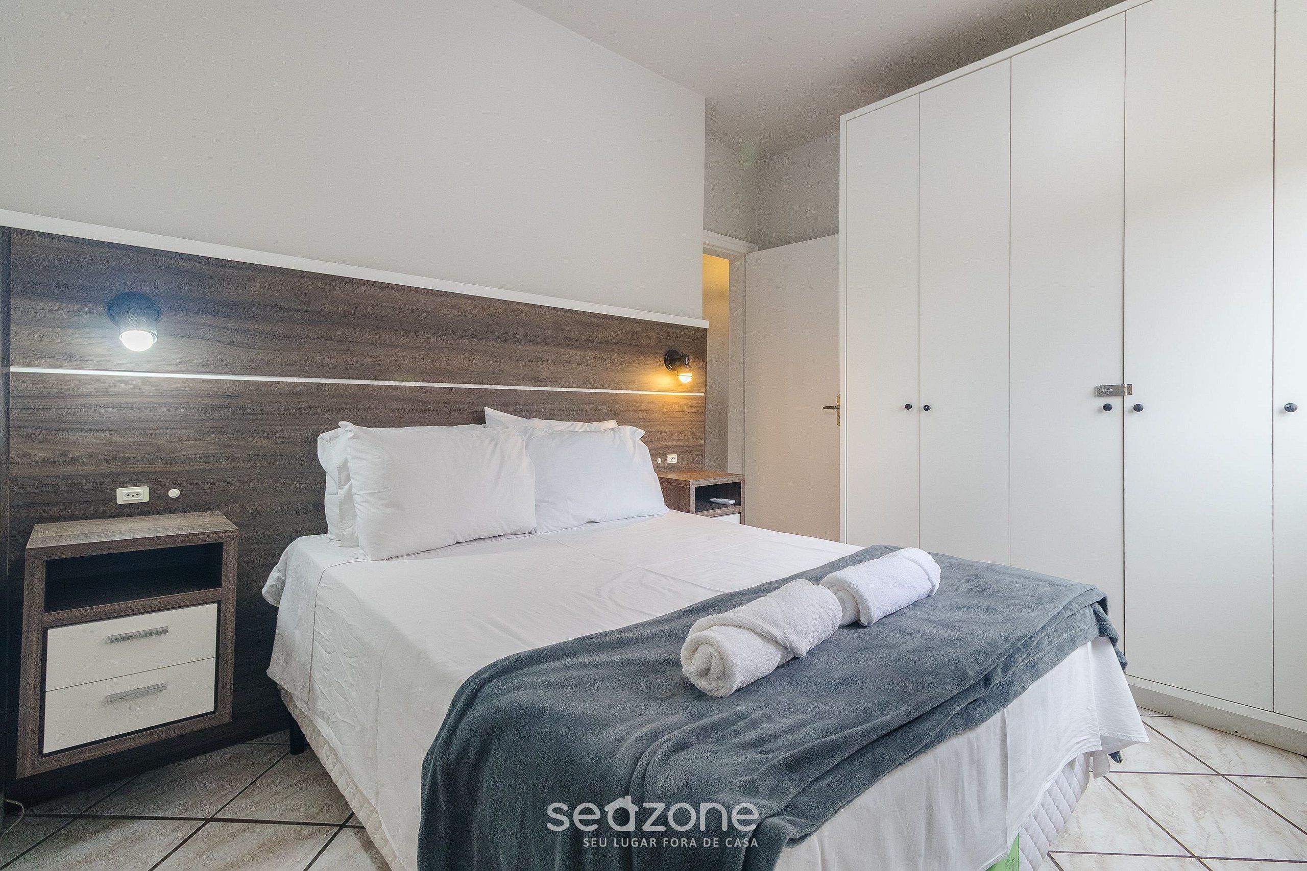 A neatly made bed with a gray blanket and a rolled towel, a bedside table with a lamp, and a wardrobe with a mirror.