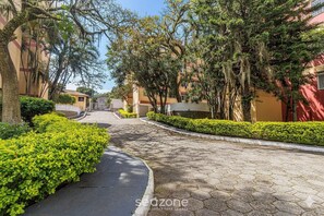 A paved pathway flanked by neatly trimmed hedges and trees, leading towards a building with a red facade. - Apartment With 2 Bedrooms in Floripa Opt0101 (Florianópolis)
