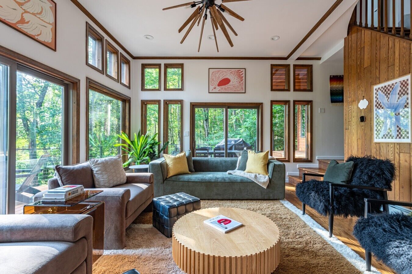 A spacious living room with large windows, a wooden ceiling fan, and a variety of seating options including a sofa, chairs, and a round coffee table.