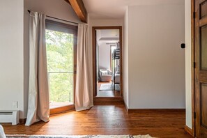 A hallway with wooden flooring, a view through a glass door, and a glimpse of a bedroom with a bed and a chair.