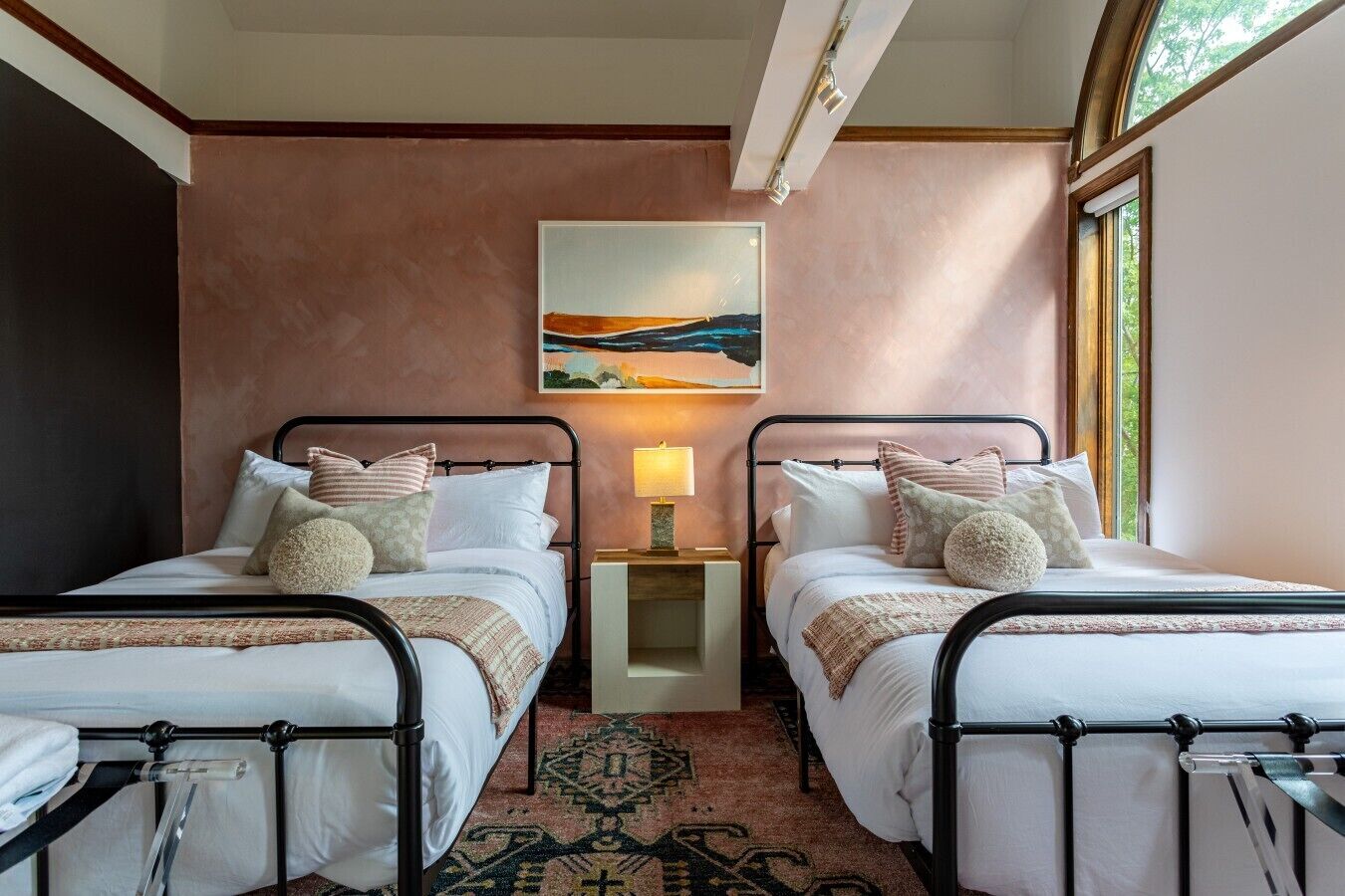 A room with two beds, a nightstand with a lamp, a painting on the wall, and a window.