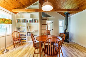 A wooden dining table with chairs, a bookshelf, a ladder, and a wood-burning stove in a room with wooden floors and a sloped ceiling.
