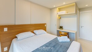 Una habitación de hotel moderna con un cabecero de madera, una cama con sábanas blancas, un plaid gris y un estante empotrado con un espejo y toallas dobladas.