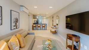 Una sala de estar moderna con un sofá, una mesa de centro y un televisor. En el fondo, una cocina con una barra y sillas.