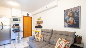 Een moderne woonkamer met een grijze bank, kleurrijke kussens met uilen, een keukenhoek met een koelkast, magnetron en waterkoker, en een deur naar een andere kamer.