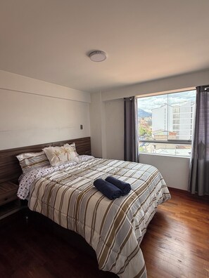 Quarto de hotel com uma cama, janela com cortinas, piso de madeira e uma luminária no teto.
