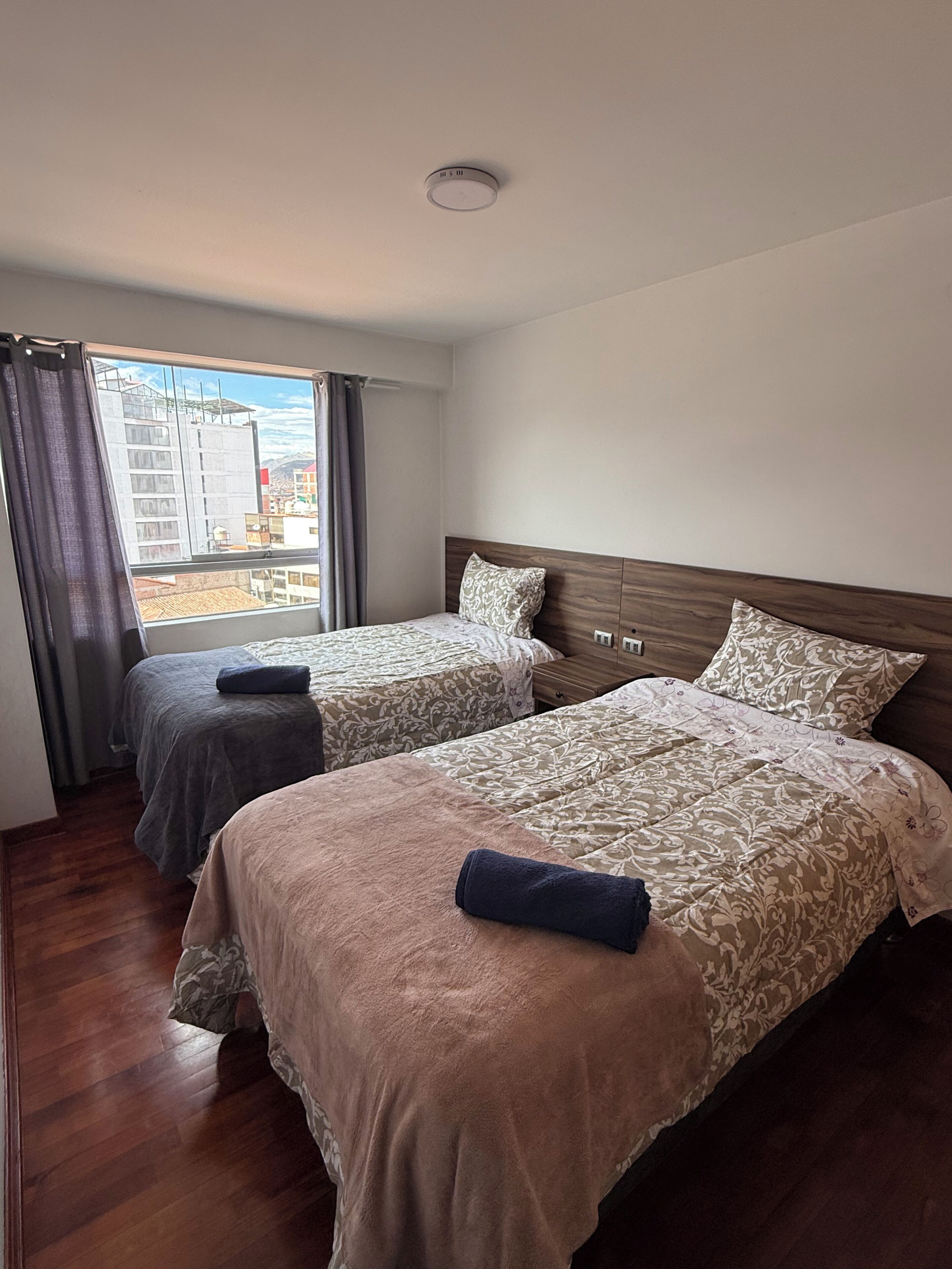 A hotel room with two beds, a window with curtains, a wooden headboard, and a view of buildings outside.