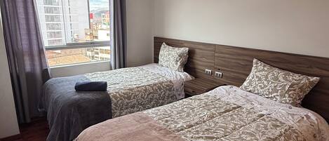 A hotel room with two beds, a window with curtains, a wooden headboard, and a view of buildings outside.