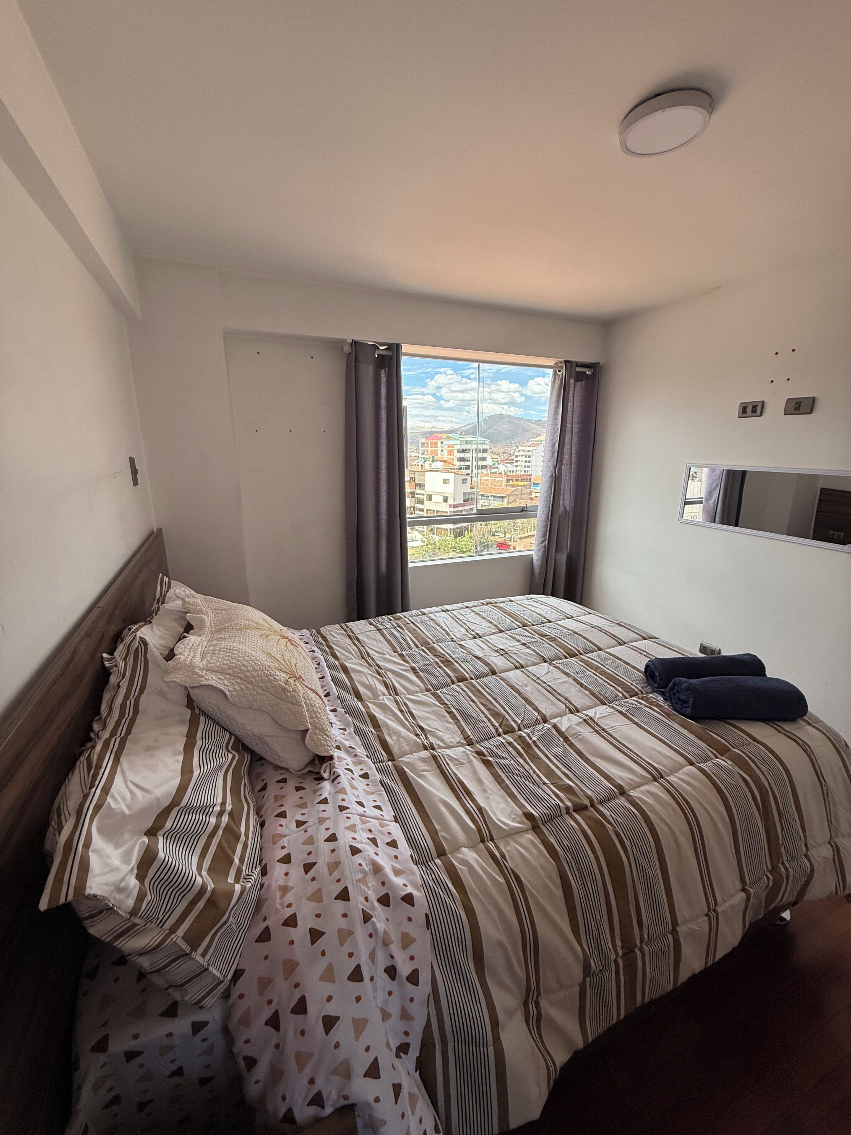 Ein Hotelzimmer mit einem Bett, einem fensterbelaubten Fenster, einem an der Wand befestigten Flachbildfernseher und Blick auf Gebäude und Berge.