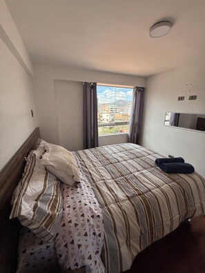 A hotel room with a bed, a window with curtains, a flat-screen TV mounted on the wall, and a view of buildings and mountains outside. - Beautiful 2-bedroom apartment perfect for a relaxing getaway. (Wanchaq)
