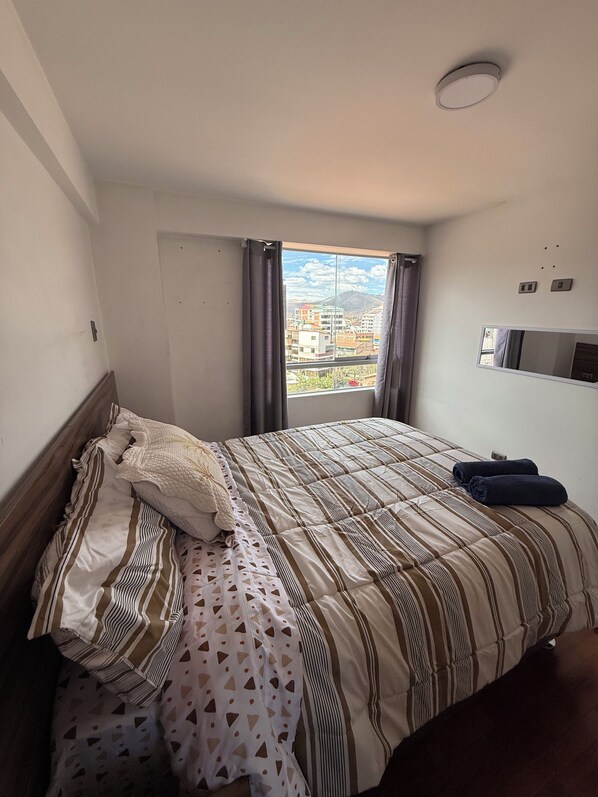 Ett hotellrum med en säng, ett fönster med gardiner, en platt-TV som är monterad på väggen och utsikt över byggnader och berg.