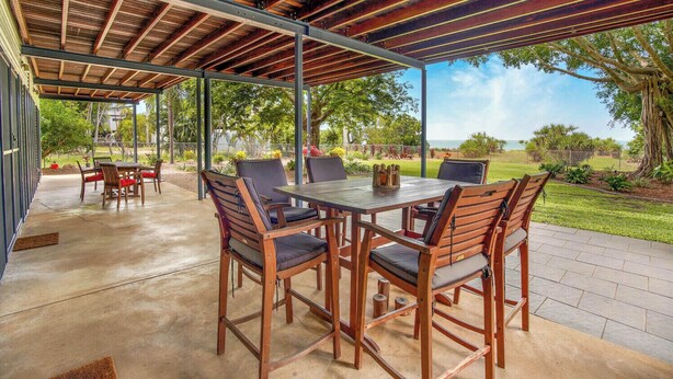 An outdoor patio with a wooden table and chairs, a covered area with a metal roof, and a view of a landscaped garden and ocean.