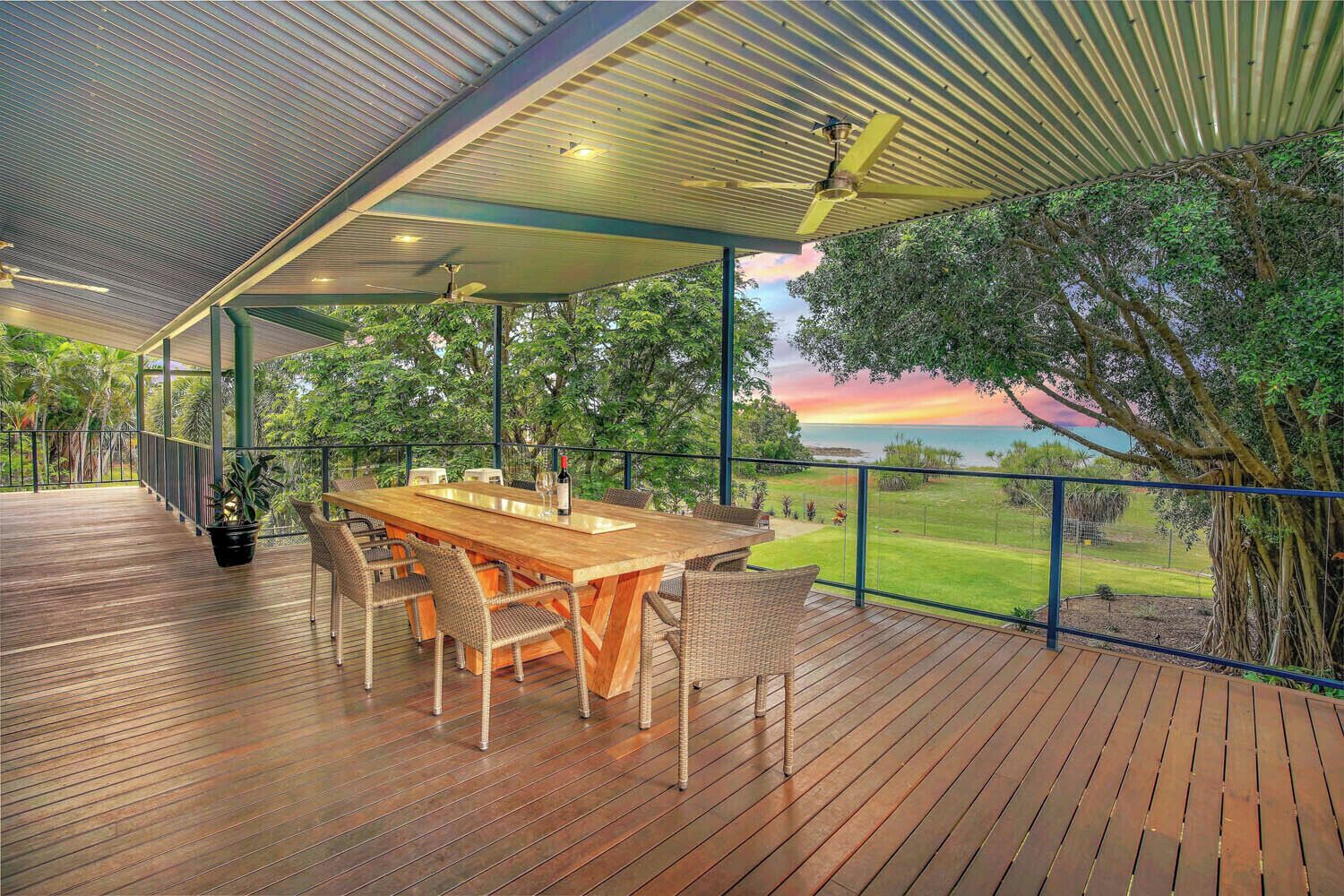 Un espace de restauration en plein air avec une table et des chaises en bois, entouré d’une terrasse en bois et d’une balustrade en verre donnant sur un paysage luxuriant.