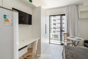 Quarto de hotel moderno com uma secretária branca, televisão de tela plana, área de refeições com mesa e cadeiras, e varanda com vista para outros edifícios.