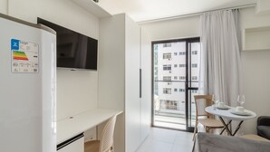 Una habitación de hotel moderna con un escritorio blanco, un televisor de pantalla plana, una zona de comedor con mesa y sillas, y un balcón con vistas a otros edificios.