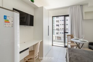 A modern hotel room with a white desk, a flat-screen TV, a dining area with a table and chairs, and a balcony with a view of other buildings. - Studio 450m From Lighthouse & Beach - Prv0404 (Salvador)