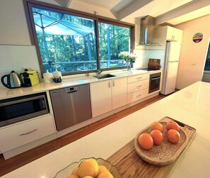 A modern kitchen with a large window, stainless steel appliances, and a wooden cutting board with oranges.